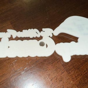 May include: White plastic ornament with the words "Happy Holidays" and "Greetings" in a festive font. The ornament has four silver metal fasteners.