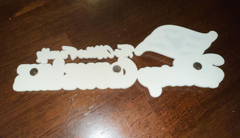 May include: White plastic ornament with the words "Happy Holidays" and "Greetings" in a festive font. The ornament has four silver metal fasteners.