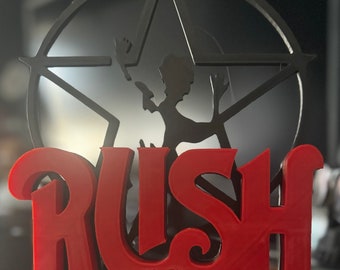 May include: A black metal 3D printed sculpture of the band Rush. The sculpture features a five-pointed star with a silhouette of a person in the center. The word "RUSH" is printed in red below the star.