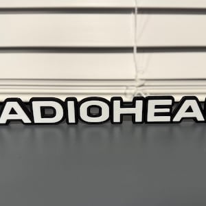 May include: A white, raised, plastic lettering spelling out the word "RADIOHEAD" with black outlines.