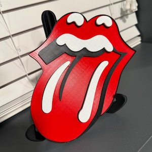 May include: A red and white 3D printed tongue with a black outline. The tongue is sticking out and has a white line down the middle. The tongue is on a black stand.