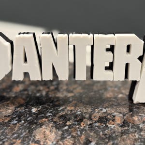 May include: White 3D printed letters spelling out the word "PANTERA" on a brown speckled surface.