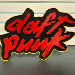 May include: A black sign with the words "DAFT PUNK" in red, outlined in yellow. The sign is mounted on a wall with a window and blinds in the background. The sign is a decorative item.
