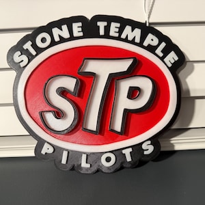 May include: A decorative sign featuring the text "STONE TEMPLE PILOTS" in a black, white, and red color scheme. The sign has an oval shape with the letters "STP" in the center.