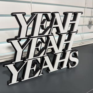 May include: A black and white wooden sign that spells out "YEAH YEAH YEAH" in a stacked design. The letters are white with black outlines, creating a 3D effect. The sign is placed on a dark gray surface.