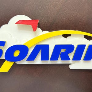 May include: A white 3D printed sign with a yellow arc and the word "SOARIN'" in blue letters. A red and white airplane is in the upper left corner of the sign.
