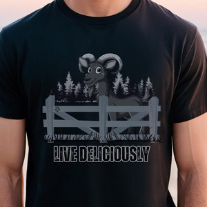 Puede incluir: Camiseta negra con un diseño de carnero de dibujos animados detrás de una valla de madera, con un bosque de fondo. El texto "LIVE DELICIOUSLY" está impreso debajo en blanco. La camiseta es de color negro liso.
