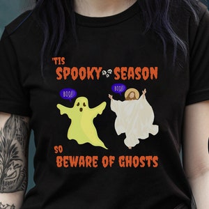 Puede incluir: Camiseta negra con un gráfico de un fantasma y Jesús con el texto "'Tis Spooky Season So Beware of Ghosts".
