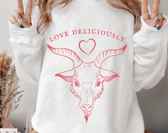 Peut inclure: Sweat-shirt blanc avec un motif rouge d'une tête de chèvre, d'un cœur et l'inscription "LOVE DELICIOUSLY". Le sweat-shirt à col rond et à manches longues présente un motif centré sur le devant.