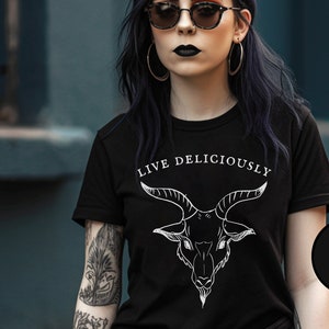 Puede incluir: Una camiseta negra de manga corta con un gráfico de líneas blancas de una cabeza de cabra centrado en el pecho. Sobre la ilustración de la cabra, el texto "LIVE DELICIOUSLY" aparece en una fuente serif blanca. Un círculo negro indica "BLACK".