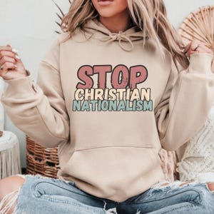 May include: A beige hoodie with the text "STOP CHRISTIAN NATIONALISM" in a retro style font. The text is in shades of pink, yellow, and blue.