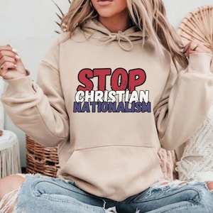 Puede incluir: Una sudadera con capucha beige con el texto "STOP CHRISTIAN NATIONALISM" impreso en rojo, blanco y azul.