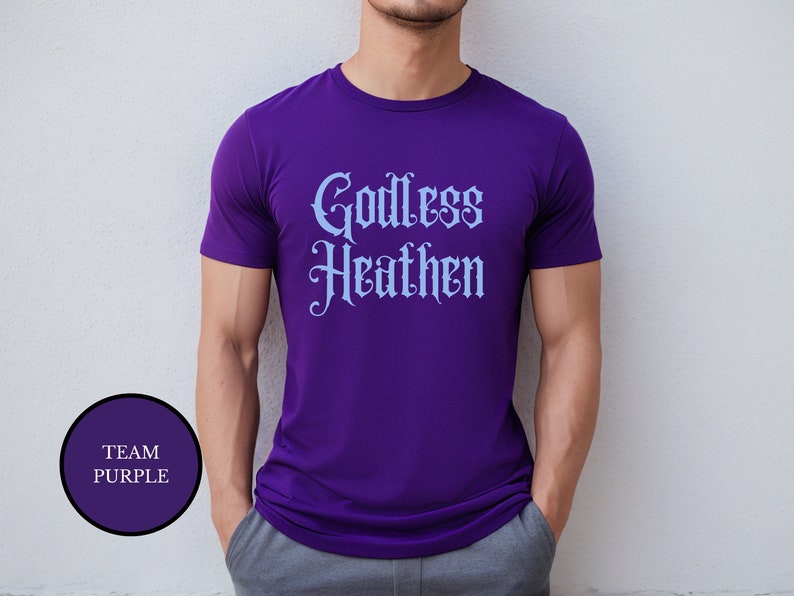 Gay man wearing an anti religion shirt that says Godless Heathen in gothic pastel blue font on a purple shirt