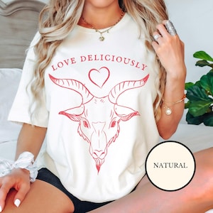 Puede incluir: Camiseta blanca con un gráfico rojo de una cabeza de cabra con un corazón encima y el texto "LOVE DELICIOUSLY". La camiseta está puesta en una persona que lleva pantalones cortos negros. El texto "NATURAL" está en un círculo en el lado derecho de la imagen.