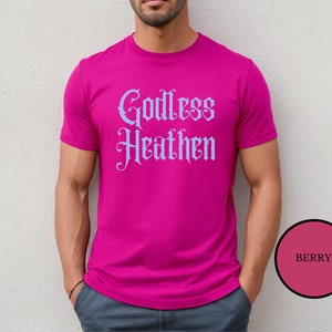 Leftist male wearing an anti religion shirt that says Godless Heathen in gothic pastel blue font on a pink shirt