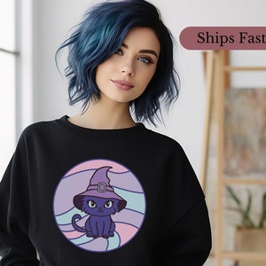 Op de afbeelding: Zwart sweatshirt met een paarse en roze afbeelding van een kat met een heksenhoed. De kat zit in een cirkel met een golvend ontwerp.