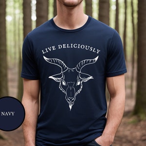 A male warlock in the woods wearing a dark navy blue witch aesthetic goth shirt featuring Black Phillip demon goat that reads Live Deliciously like a proud witch
