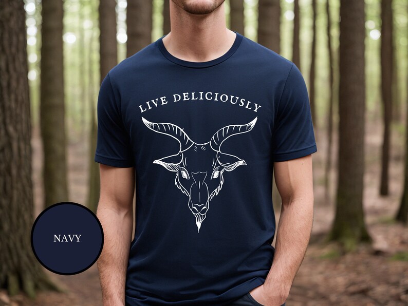 A male warlock in the woods wearing a dark navy blue witch aesthetic goth shirt featuring Black Phillip demon goat that reads Live Deliciously like a proud witch