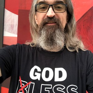 White male hipster with long graying hair wearing a black atheist shirt that reads God Bless across the front, but the B on Bless has a red X so the message reads GODLESS. This Atheist shirts contrasts against the red artwork in the background.