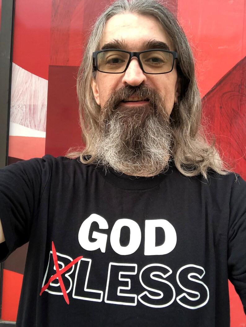White male hipster with long graying hair wearing a black atheist shirt that reads God Bless across the front, but the B on Bless has a red X so the message reads GODLESS. This Atheist shirts contrasts against the red artwork in the background.