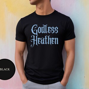 Gay male against a rainbow painted wall wearing an anti religion shirt that says Godless Heathen in gothic pastel blue font on a snug black shirt that clings to his sculpted muscles