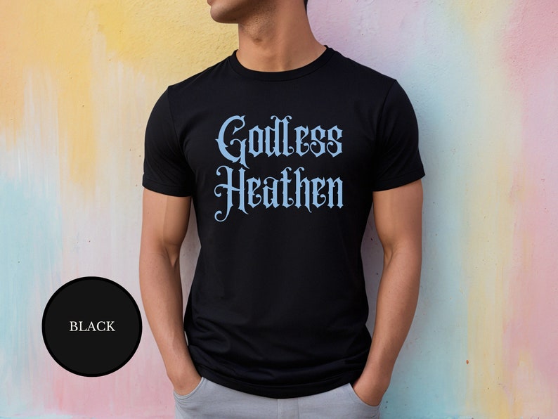 Gay male against a rainbow painted wall wearing an anti religion shirt that says Godless Heathen in gothic pastel blue font on a snug black shirt that clings to his sculpted muscles