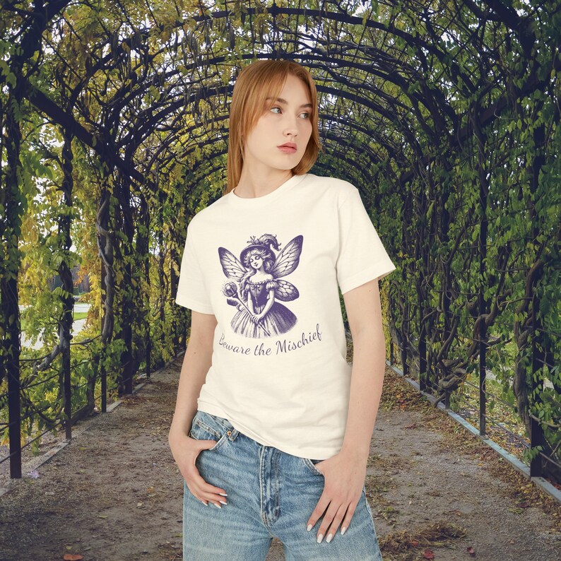 Redhead Gen Z girl wearing a Unisex Faded Cream Vintage Style Retro Fairy Grunge T-Shirt featuring a Purple Winged Fairy and the sarcastic saying Beware the Mischief on this cute whimsical nature shirt for fairy lovers, as she walks through a botanical garden.