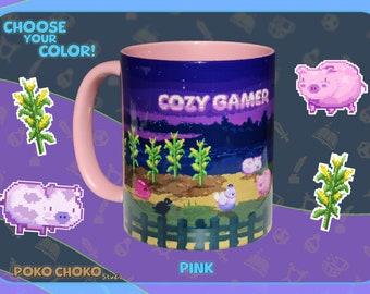 May include: Pink ceramic mug with a pixelated design of a farm scene at night. The mug says "Cozy Gamer" and has a pink handle. The mug is surrounded by pixelated images of corn, pigs, and a chicken.