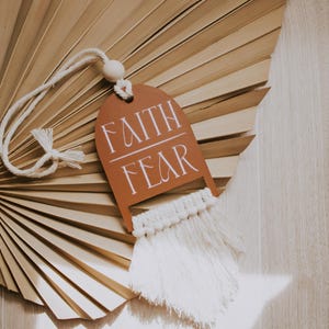 May include: A decorative tag with the words "FAITH FEAR" in white script, hanging from a white cord with a wooden bead. The tag is brown and has a macrame fringe. The background features a dried palm leaf fan.