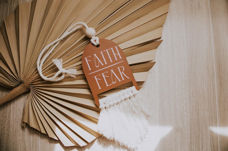 May include: A decorative tag with the words "FAITH FEAR" in white script, hanging from a white cord with a wooden bead. The tag is brown and has a macrame fringe. The background features a dried palm leaf fan.