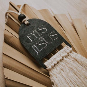 May include: A dark gray wooden wall hanging with the text "EYES on JESUS" in white script. It features a macrame fringe and a black wooden bead. The background is a dried palm leaf.