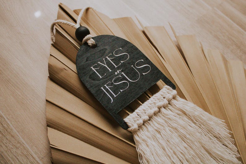 May include: A dark gray wooden wall hanging with the text "EYES on JESUS" in white script. It features a macrame fringe and a black wooden bead. The background is a dried palm leaf.
