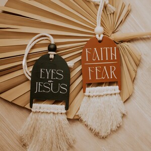 May include: Two decorative wooden tags with macrame fringe. One tag is dark green with "EYES on JESUS" in white. The other is rust-colored with "FAITH FEAR" in white. Both have a cream-colored rope for hanging.