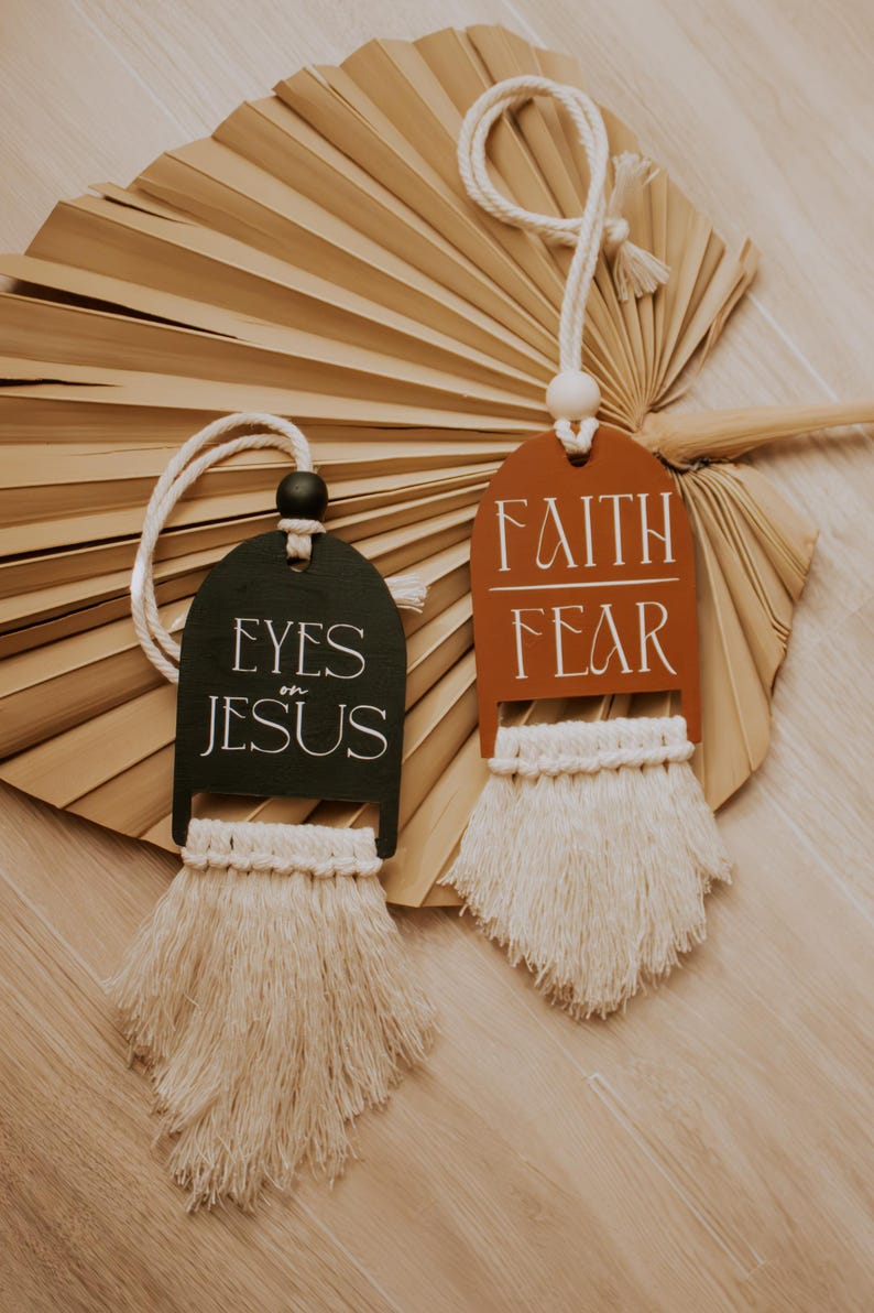 May include: Two decorative wooden tags with macrame fringe. One tag is dark green with "EYES on JESUS" in white. The other is rust-colored with "FAITH FEAR" in white. Both have a cream-colored rope for hanging.