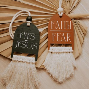 May include: Two decorative wooden hangings with macrame fringe. One is dark green with the words "EYES on JESUS" in white. The other is rust-colored with "FAITH FEAR" in white. Both have a cream-colored cord for hanging.