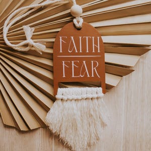 May include: A brown wooden tag with the words "FAITH" and "FEAR" in white script, hanging from a white rope. The tag has a macrame fringe at the bottom. The background features a dried palm leaf.