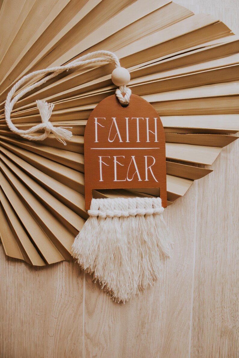 May include: A brown wooden tag with the words "FAITH" and "FEAR" in white script, hanging from a white rope. The tag has a macrame fringe at the bottom. The background features a dried palm leaf.