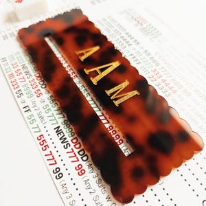 May include: A rectangular tortoiseshell pattern mahjong tile rack with scalloped edges and the gold embossed letters AAM centered on the front. The rack is positioned over a printed mahjong score card with red and black text.