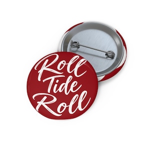 May include: A round pin-back button featuring a deep red background with the white script text Roll Tide Roll printed across the front. The back of the button is silver-toned metal and includes a horizontal pin mechanism for attachment to clothing.