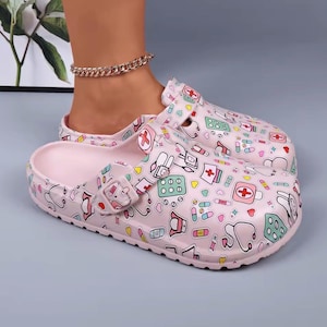 May include: Pink clogs with a medical-themed pattern featuring stethoscopes, pills, and first aid symbols. The clogs have a buckle closure and a molded footbed. The shoes are designed for comfort and support.