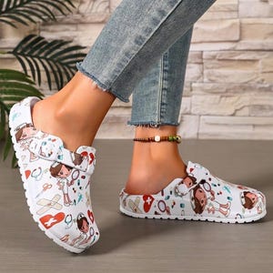 May include: White slip-on clogs featuring a colorful medical-themed print. The pattern includes cartoon nurses, red hearts, stethoscopes, scissors, bandages, and various pills. Each shoe has an adjustable strap across the top with a buckle detail.