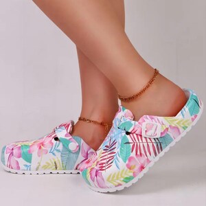 May include: White clogs with a colorful floral pattern featuring pink, blue, and green leaves and flowers. The clogs have a buckle closure and a white sole. The overall theme is a tropical or summer style.