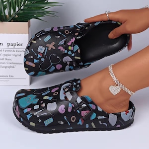 May include: Black clogs with a medical-themed pattern featuring stethoscopes, pills, and bandages. The clogs have a buckle closure and a molded footbed. The design includes blue, pink, and white accents.