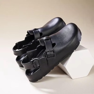 May include: A pair of black clogs with adjustable straps and buckle closures. The clogs are made of a smooth material and have a contoured footbed. The shoes are displayed on a white geometric shape.