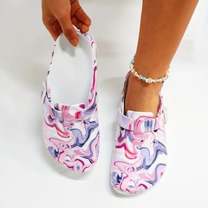 May include: White clogs with a pink and purple marble pattern. The clogs have a buckle strap and a white sole.