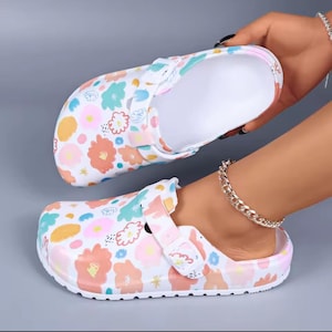 May include: White clogs with a colorful floral pattern in shades of pink, orange, blue, and green. The clogs have a white interior and a strap for a secure fit. These casual shoes are perfect for everyday wear.