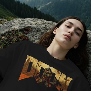 May include: Black t-shirt featuring the word "DOOM" in a pixelated, orange and gold design. The graphic includes a monster and bats. The shirt is worn by a person lying on a rock with a mountain backdrop.