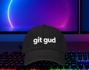 May include: Black baseball cap with the white text "git gud" across the front. The cap is sitting on a desk in front of a computer keyboard and monitor. The background has a blue and purple gradient.
