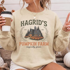May include: A beige crewneck sweatshirt featuring a graphic print of a cabin surrounded by pumpkins. The text reads HAGRID'S EST. 1981 PUMPKIN FARM magically grown. The design includes green lettering, a dark cabin, orange pumpkins, and leafy branches.