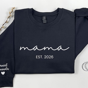May include: A navy blue sweatshirt with the word "mama" in white script, and "EST. 2026" below it. The left sleeve has the names "Daniel Amanda" and a heart. Two silver hoop earrings are on the right. A sprig of dried flowers is on the left.